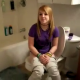 A blonde girl sits on the toilet sideways as her friend observes. We think she is taking a shit, but she does not say.